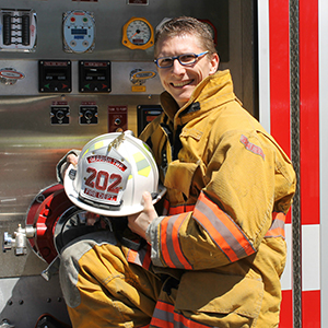 Assistant Fire Chief, Brandon Rodgers, posing in front of a Gerrish Township fire engine in his bunker gear and holding his helmet.